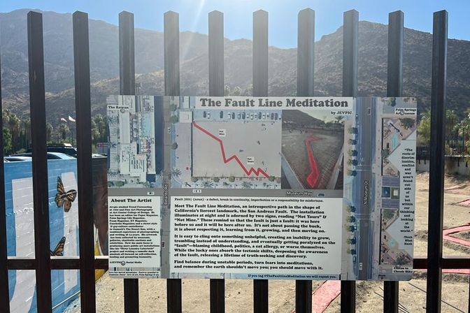 Interpretive sign titled The Fault Line Meditation for a fault-line art installation, mounted on a metal fence with a red zigzag path diagram and desert mountains under a sunny blue sky.
