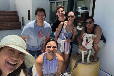Smiling group of six friends in casual summer clothes with a happy white dog (tongue out) posing for a selfie on a sunny urban sidewalk outside a building.