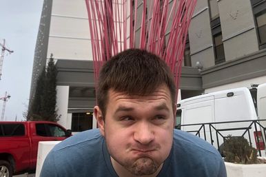 Person puffing cheeks playfully in front of a tall red metal sculpture outside an urban building, with parked vans and a red pickup truck nearby.