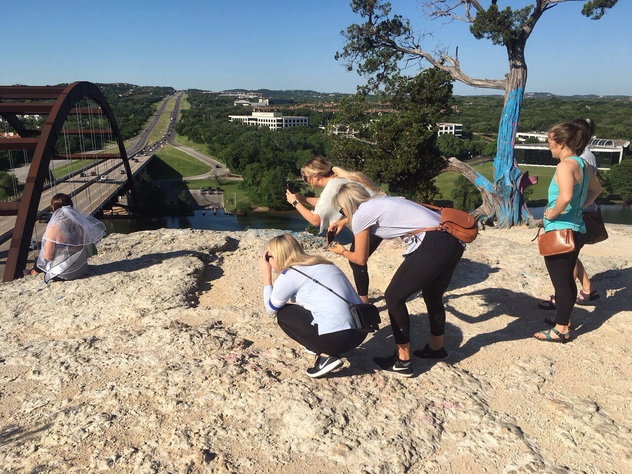Group of women photographing a veiled woman on a rocky Austin, Texas overlook above the Pennybacker (360) Bridge and highway, sunny day with a painted tree in the background