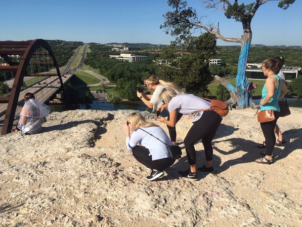 Group of women photographing a veiled woman on a rocky Austin, Texas overlook above the Pennybacker (360) Bridge and highway, sunny day with a painted tree in the background