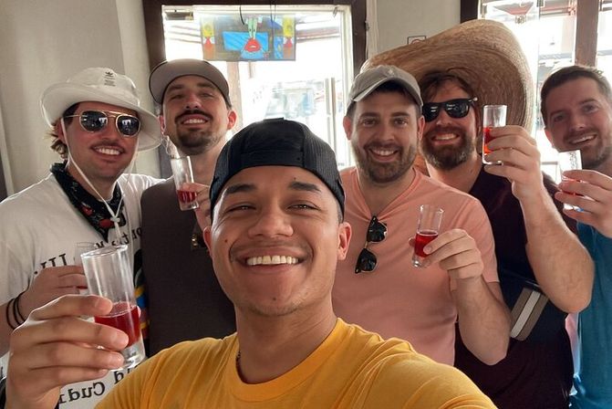 Six friends taking a selfie while toasting with small red cocktails in a bright bar, wearing caps, sunglasses and a large straw hat — casual daytime celebration.