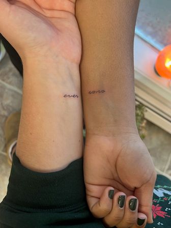 Close-up of two wrists, light and dark skin tones, showing matching tiny black cursive "ever" wrist tattoos on inner wrists against a cozy indoor background