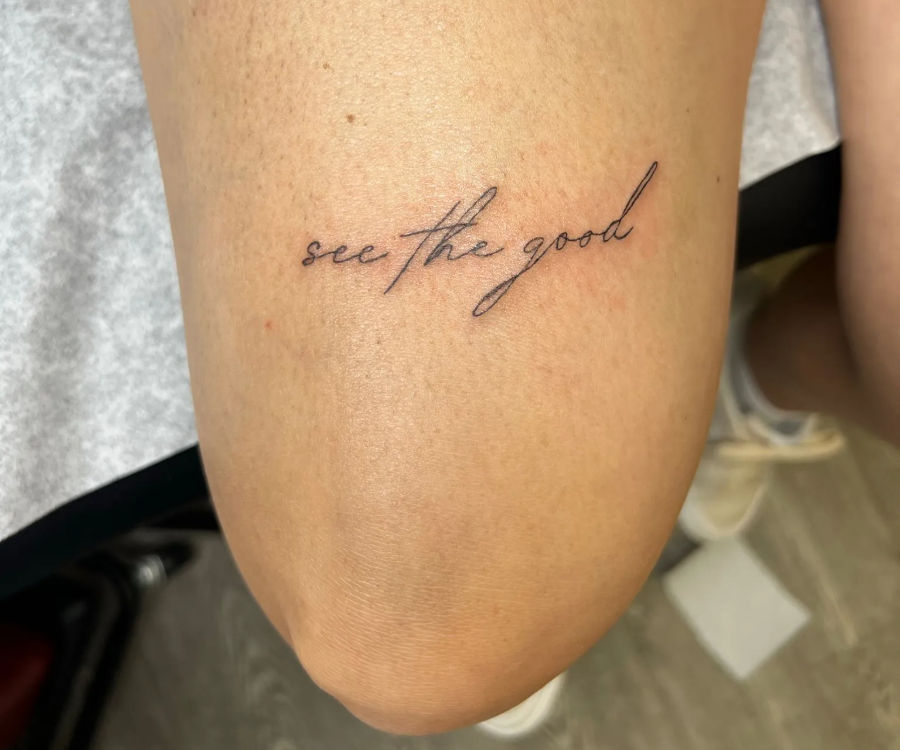 Close-up of a minimalist black-ink script tattoo reading 'see the good' on the inner knee/upper calf.