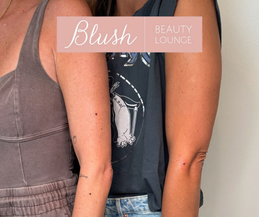 Close-up of two sun-kissed arms side by side in casual tank tops and denim, showing tiny minimalist tattoos (small heart and outline) against a neutral wall background.