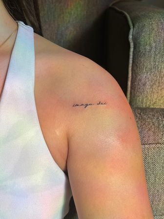 Close-up of a tiny minimalist cursive shoulder tattoo reading "imago dei" in black ink on an upper shoulder next to a white tank top.