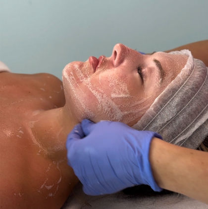 Close-up of a woman receiving an exfoliating facial at a spa — white scrub on face and neck, esthetician’s gloved hands massaging under the jaw, disposable headband in place.