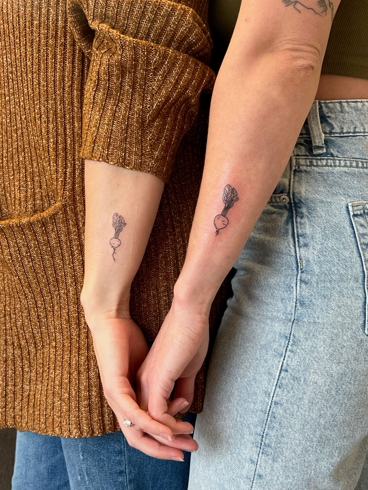 Matching minimal line-art radish tattoos on two inner forearms, hands gently clasped — one person wearing a brown ribbed sweater, the other in light denim jeans.