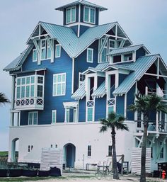 Blue coastal beach house with white trim and multiple metal gabled roofs, wraparound balconies, palm trees and construction materials on a sandy lot