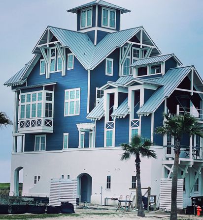 Blue coastal beach house with white trim and multiple metal gabled roofs, wraparound balconies, palm trees and construction materials on a sandy lot