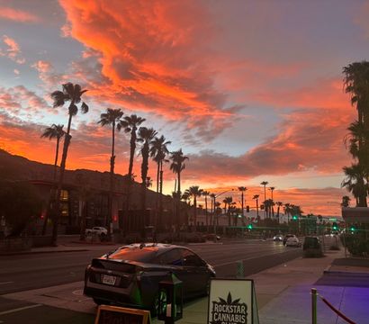 Vibrant orange-pink sunset over a palm-lined main street with silhouetted mountains, parked cars, neon storefront lights, and glowing streetlights in a desert-city scene.