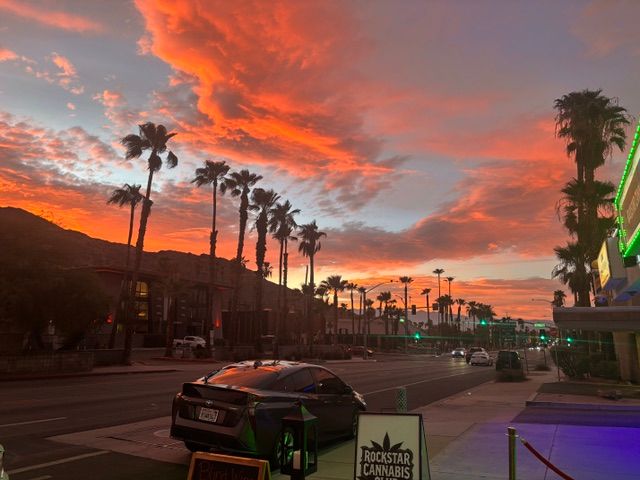 Vibrant orange-pink sunset over a palm-lined main street with silhouetted mountains, parked cars, neon storefront lights, and glowing streetlights in a desert-city scene.