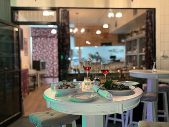 Round white table set for two with two glasses of rosé and plates of salads in a cozy indoor bistro, open kitchen and warm pendant lights glowing in the background.