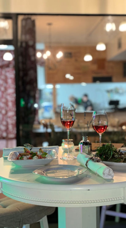 Round white table set for two with two glasses of rosé and plates of salads in a cozy indoor bistro, open kitchen and warm pendant lights glowing in the background.