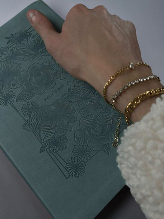 Hand wearing gold chain and crystal bracelets resting on a teal, floral-embossed Holy Bible cover with a cozy cream sleeve visible.