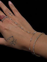 Close-up of a hand with small constellation tattoos and a French-tipped nail, styled with layered delicate beaded chain bracelets, a rhinestone bracelet and a thin ring against a black background.