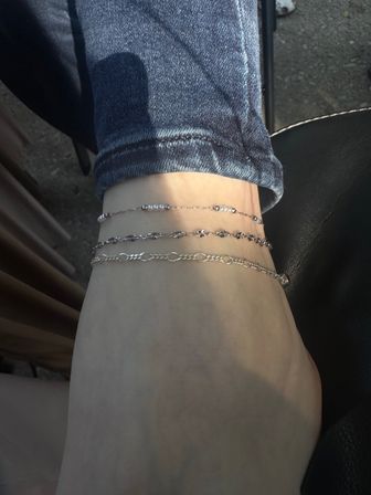 Close-up of ankle wearing three delicate silver anklets with rolled denim cuff, sunlit outdoor concrete background.