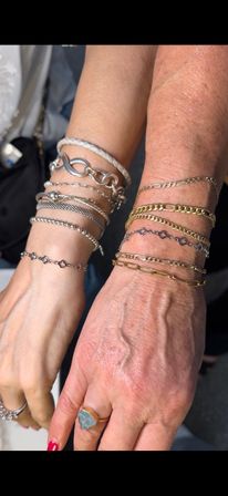 Close-up of two wrists showcasing playful stacked bracelets in mixed metals — gold and silver chains, mesh and beaded strands — paired with a rough blue gemstone ring.
