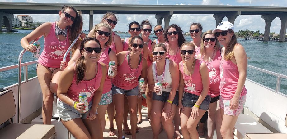 Bachelorette boat party: women in matching pink tank tops and sunglasses smiling on a sunny boat, holding canned drinks with a concrete bridge and tropical waterfront in the background.