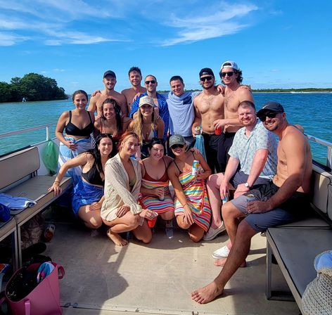 Group of friends on a sunny boat deck — about 15 young adults in swimsuits and summer outfits posing with colorful towels and drinks against a bright blue sky and calm coastal waters.