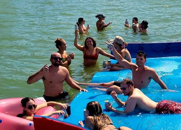 Group of friends enjoying a summer lake float on a bright blue inflatable raft, lounging in waist‑deep water and raising canned drinks