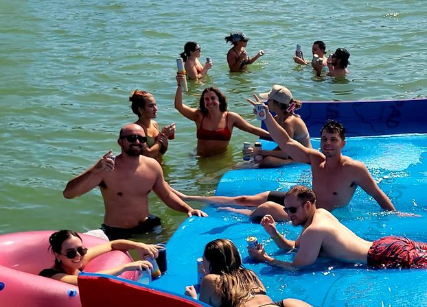 Group of friends enjoying a summer lake float on a bright blue inflatable raft, lounging in waist‑deep water and raising canned drinks