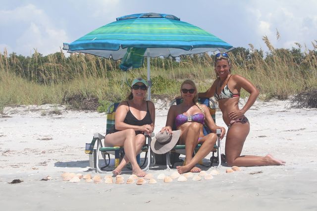 Three women relaxing under a blue-green beach umbrella on a sandy shore with dune grasses behind, two seated in beach chairs and one kneeling, smiling with a neat row of seashells arranged in the sand