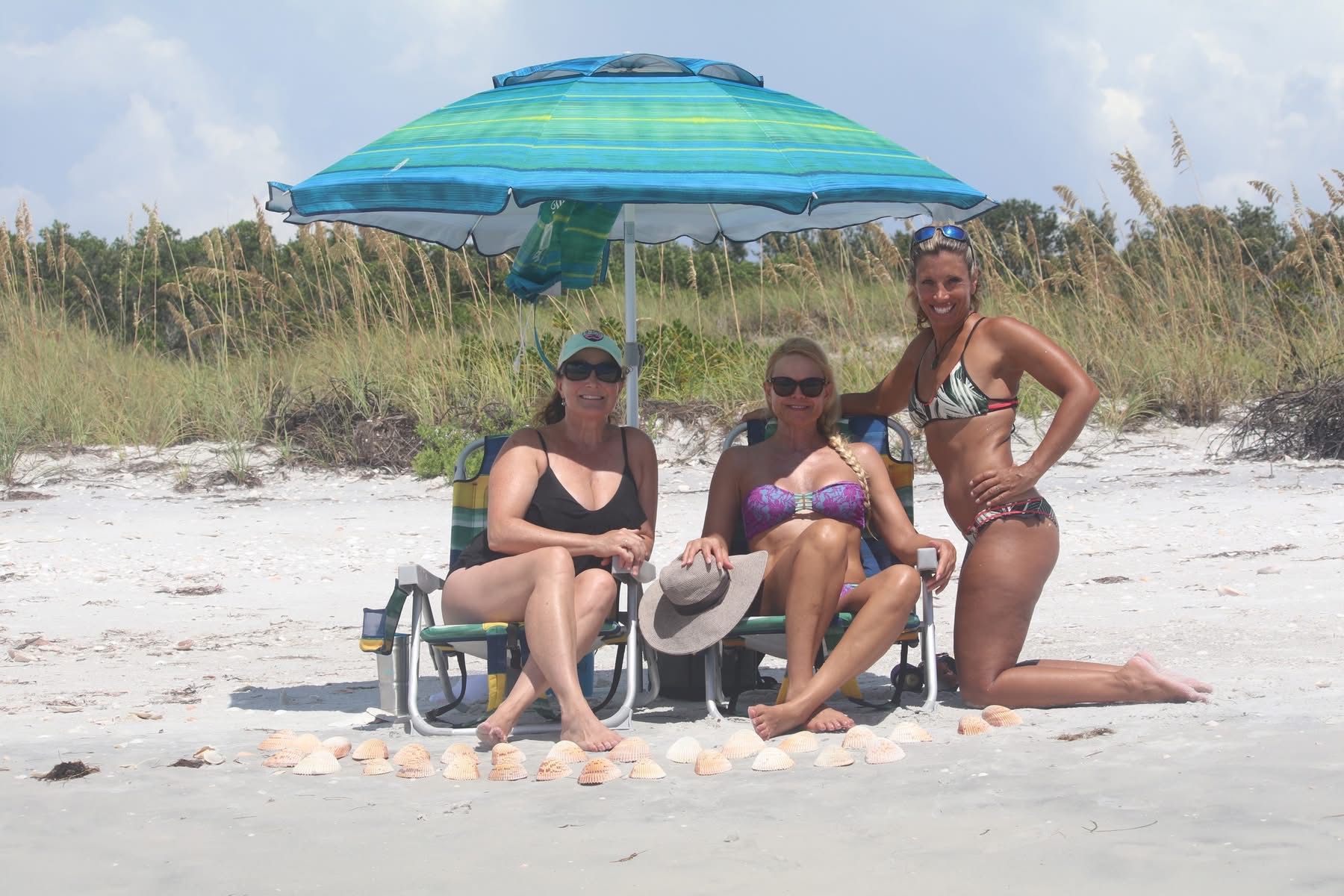 Three women in swimsuits relaxing on a sandy beach under a teal-striped umbrella with beach chairs and a neat row of seashells in front, grassy dunes behind.