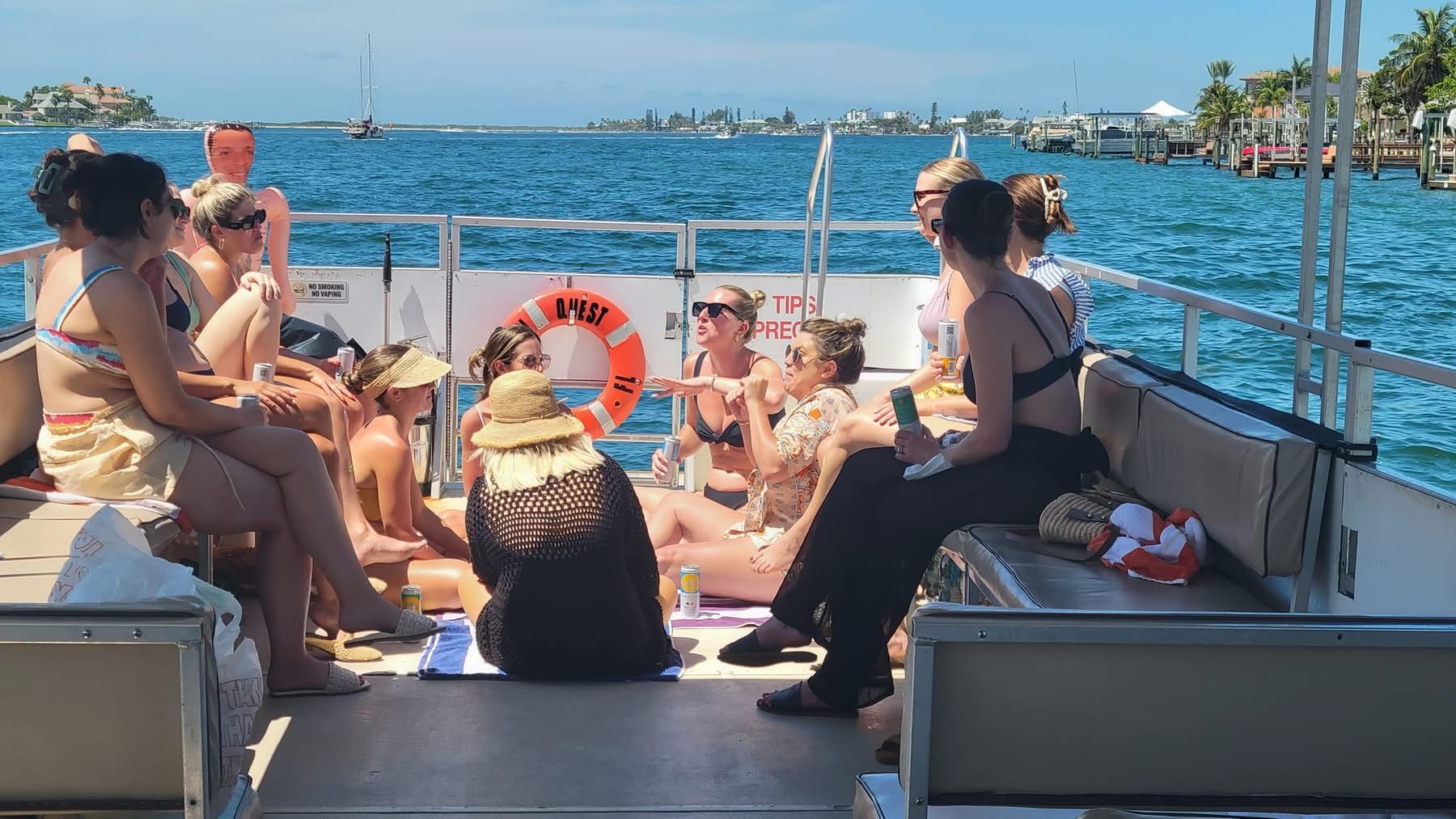 Group of women in swimsuits relaxing and chatting on a pontoon boat, sun hats and drinks, cruising a sunny coastal bay with docks and sailboats in the background.