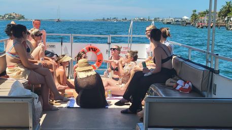 Group of women in swimsuits relaxing and chatting on a pontoon boat, sun hats and drinks, cruising a sunny coastal bay with docks and sailboats in the background.