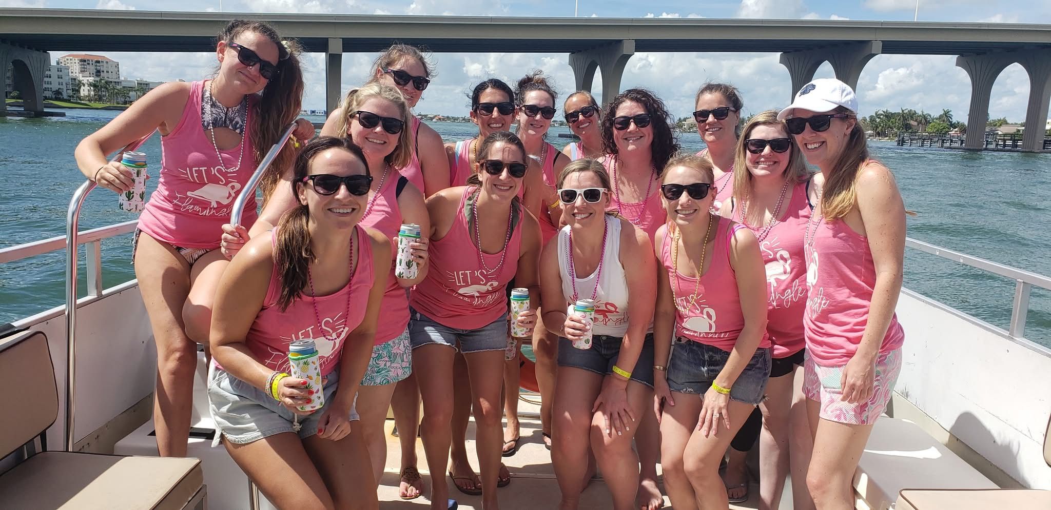 Group of women in matching pink tank tops and sunglasses smiling and holding canned drinks on a sunny boat cruise under a large concrete bridge with waterfront buildings in the background