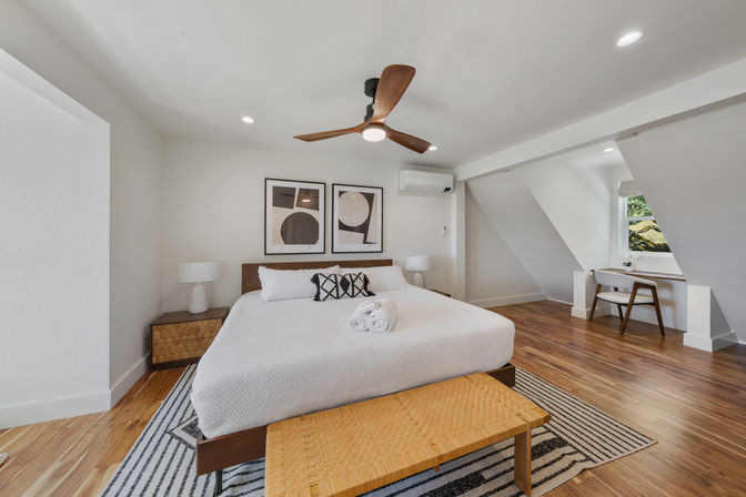 Airy modern bedroom with mid-century wooden ceiling fan, white quilted bed topped with rolled towels, abstract framed art, bedside lamps, hardwood floors and a small desk nook by a window.