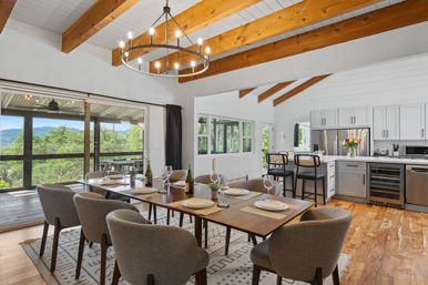 Open-concept dining room and kitchen with exposed wooden beams and circular chandelier, sliding glass doors framing lush hillside views, set dining table and breakfast island with bar stools.