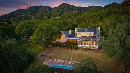 Rural hillside home with rooftop solar panels and warm glowing windows, overlooking a lit deck with lounge chairs and a swimming pool at sunset amid oak‑covered green hills.