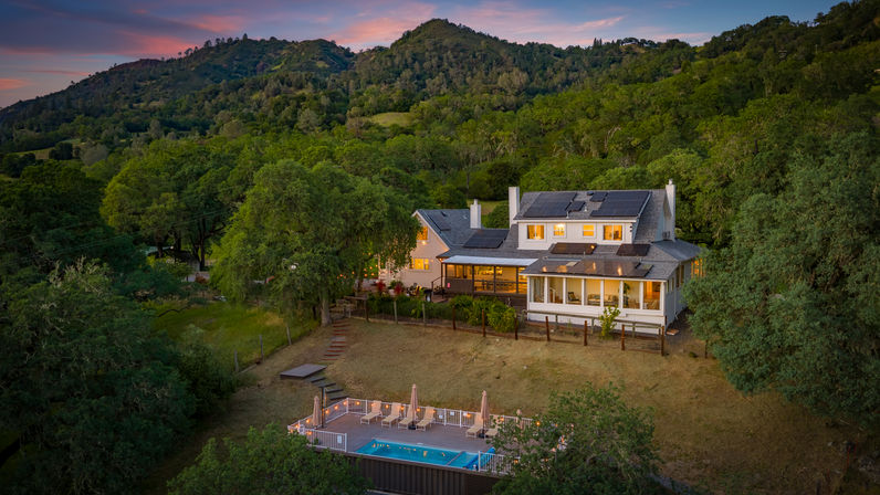 Rural hillside home with rooftop solar panels and warm glowing windows, overlooking a lit deck with lounge chairs and a swimming pool at sunset amid oak‑covered green hills.
