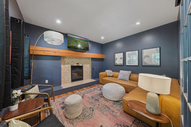 Cozy residential living room with navy blue walls, tan leather sectional, stone fireplace with wood mantel, wall-mounted TV, knit poufs, patterned boho rug, floor lamp and framed art