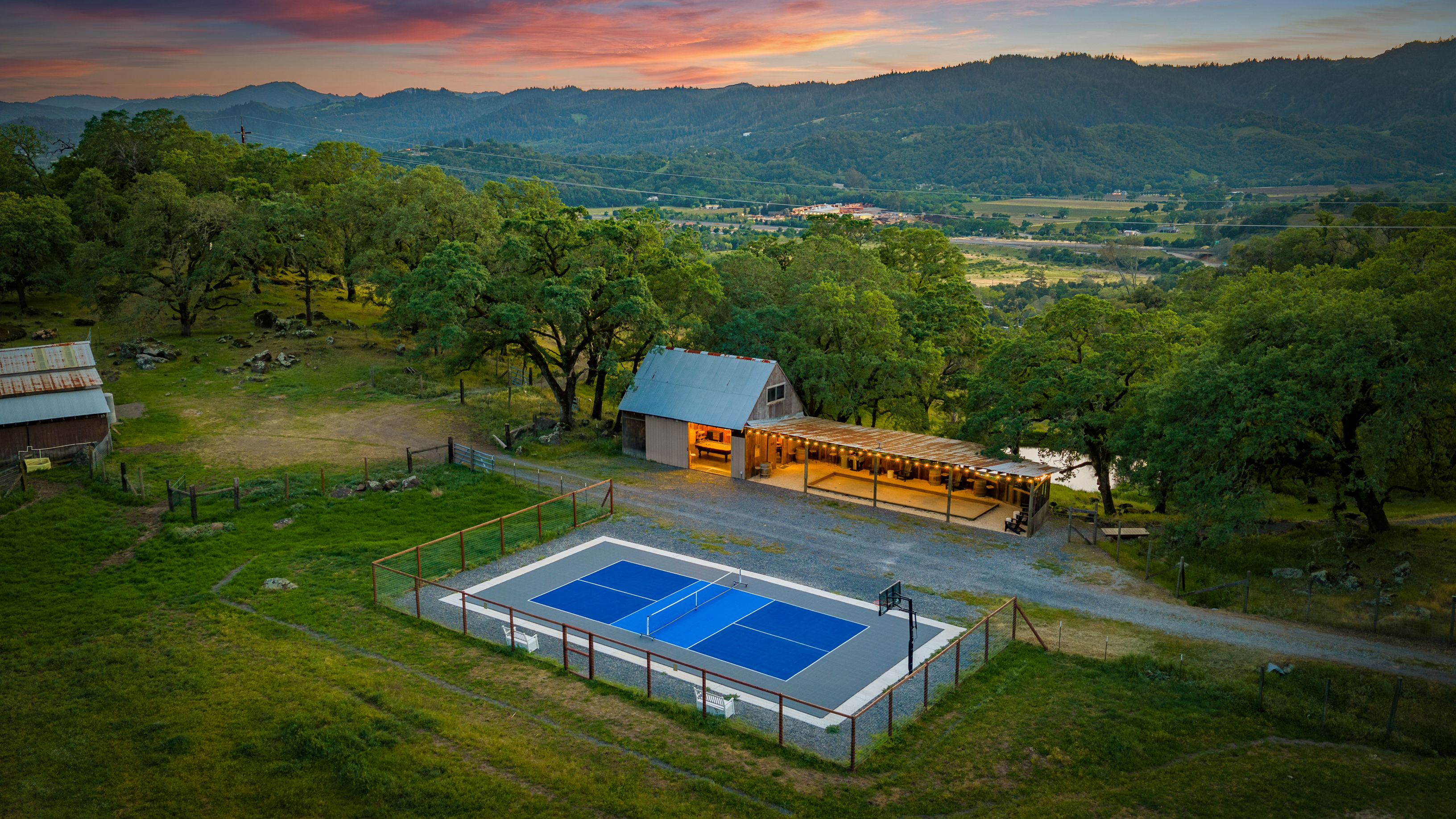 Blue outdoor tennis court fenced on a grassy hilltop beside a lit rustic barn and covered patio, surrounded by oak trees with rolling valley and mountain views under a colorful sunset.