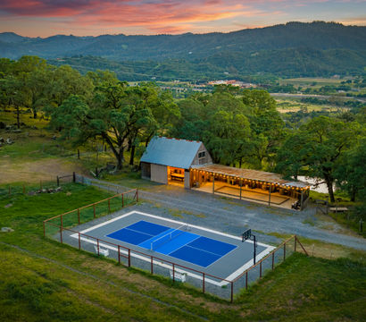 Blue outdoor tennis court fenced on a grassy hilltop beside a lit rustic barn and covered patio, surrounded by oak trees with rolling valley and mountain views under a colorful sunset.