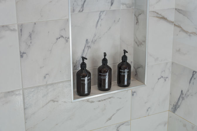Marble-tiled shower niche with three amber pump bottles on a recessed shelf — minimalist bathroom soap and shampoo display