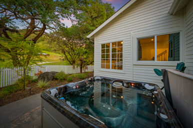 Cozy backyard hot tub on a patio beside a white clapboard house, warm interior lights visible through windows, white picket fence, mature oak trees and rolling green hills at sunset.