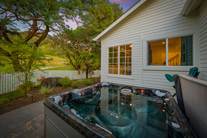 Cozy backyard hot tub on a patio beside a white clapboard house, warm interior lights visible through windows, white picket fence, mature oak trees and rolling green hills at sunset.