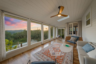 Cozy sunroom with wood floors, patterned rug, ceiling fan and skylight, panoramic windows framing a colorful sunset over rolling green hills and a backyard pool.