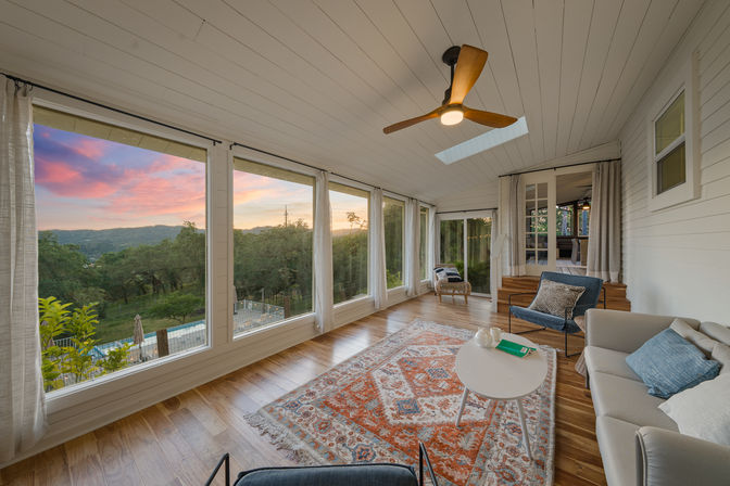 Cozy sunroom with wood floors, patterned rug, ceiling fan and skylight, panoramic windows framing a colorful sunset over rolling green hills and a backyard pool.