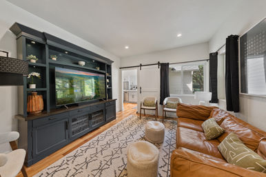 Cozy modern living room with tan leather sofa, patterned area rug, dark built-in media cabinet and TV, two accent chairs and sliding barn door to the kitchen