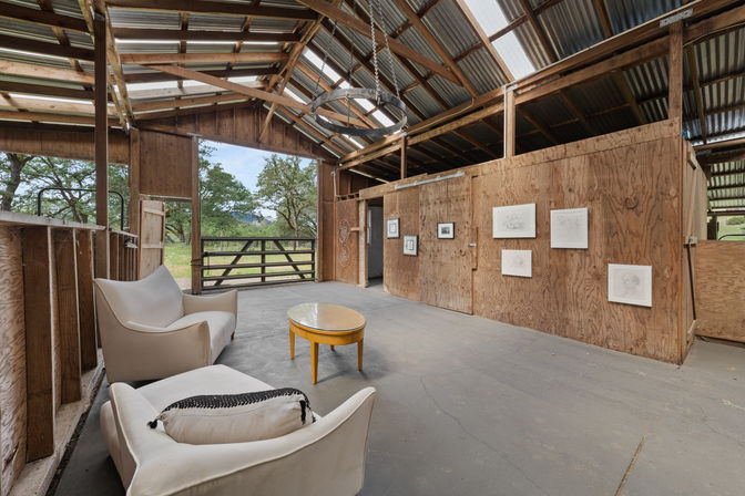 Rustic barn interior with exposed wood beams and corrugated metal roof, cozy white armchairs and a yellow round table, plywood walls hung with framed sketches, open doorway to oak-studded countryside.