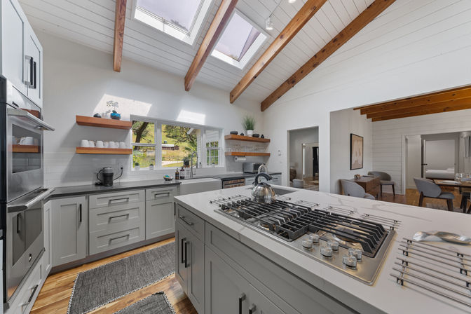 Bright modern farmhouse kitchen with skylights and exposed wooden beams, white shaker cabinets, open wood shelves, farmhouse sink, stainless appliances, large island with gas cooktop and hardwood floors