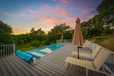 Scenic sunset over a hillside outdoor pool deck with beige umbrellas and lounge chairs, pool reflecting pink-orange sky and surrounded by oak trees and rolling green hills.