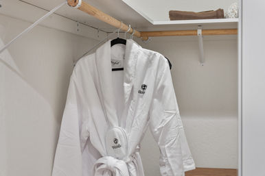 Hotel-style plush white bathrobe and matching slippers hanging on a wooden closet rod beneath a shelf with a folded towel