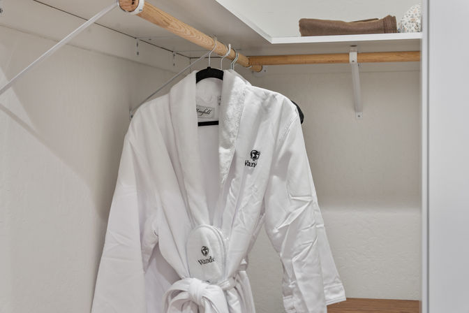 Hotel-style plush white bathrobe and matching slippers hanging on a wooden closet rod beneath a shelf with a folded towel