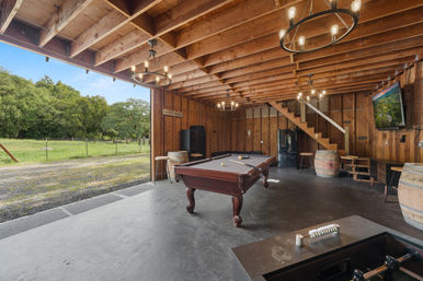 Rustic open-sided barn game room with a pool table, barrel stools, hanging chandeliers, wall-mounted TV and a wide countryside pasture view